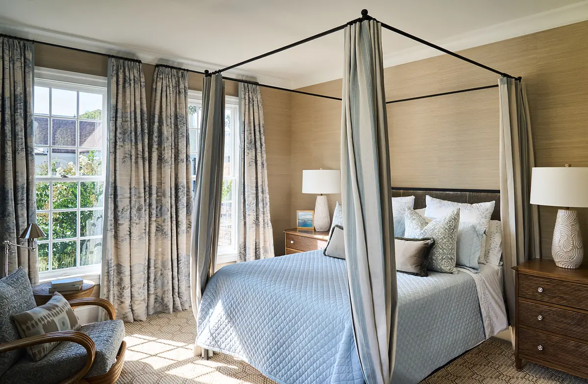 Bedroom with canopy bed, blue quilt, nightstands, table lamps, patterned curtains, and area rug.