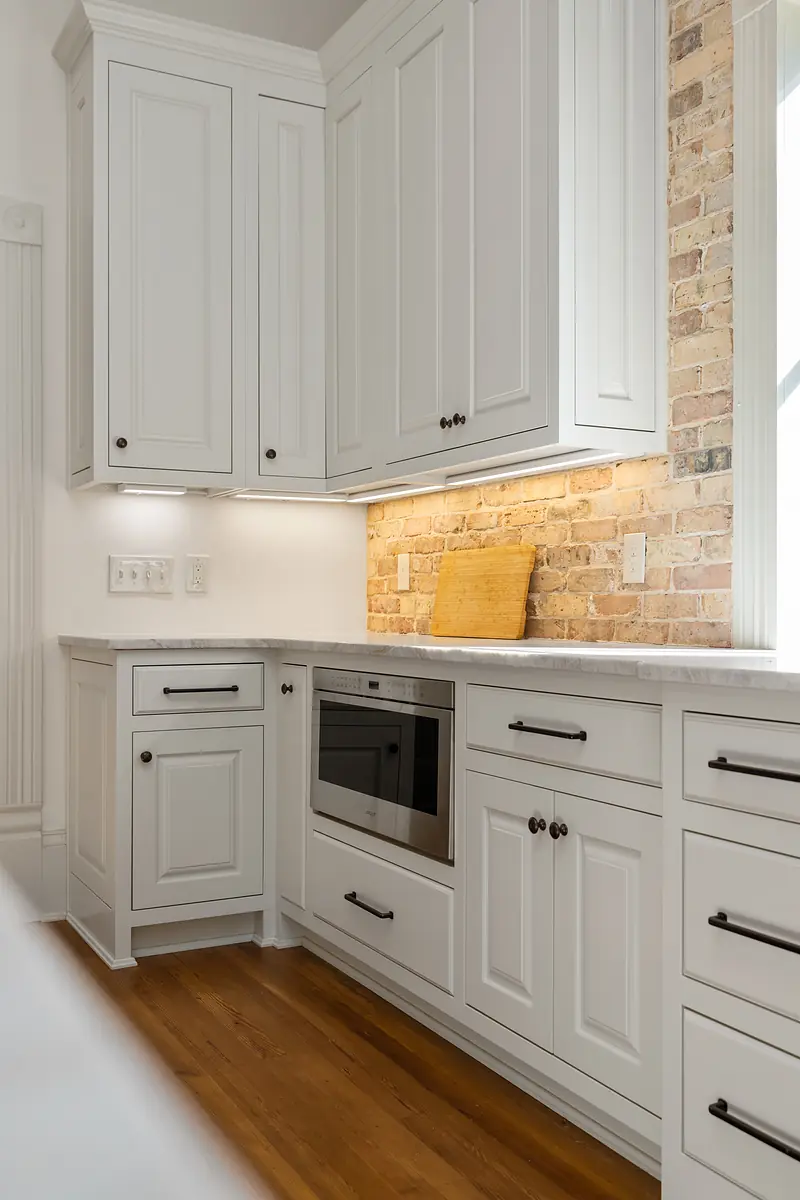 Kitchen with white cabinets, built-in microwave, wooden cutting board, brick backsplash, and hardwood floor.