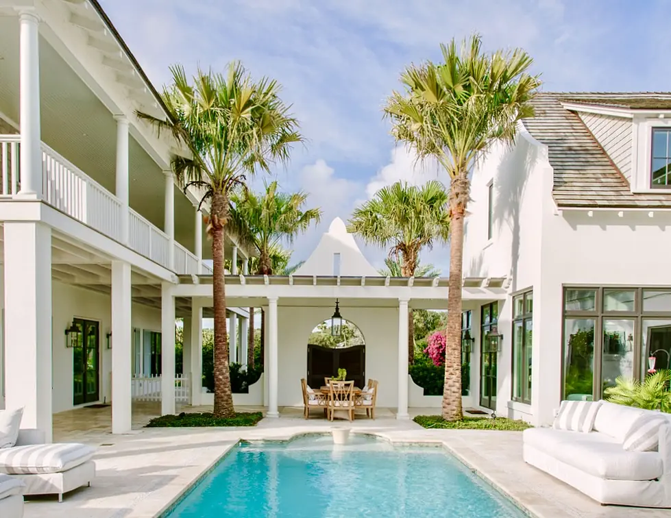 Outdoor patio with swimming pool, lounge chairs, dining table, and palm trees surrounded by white walls.