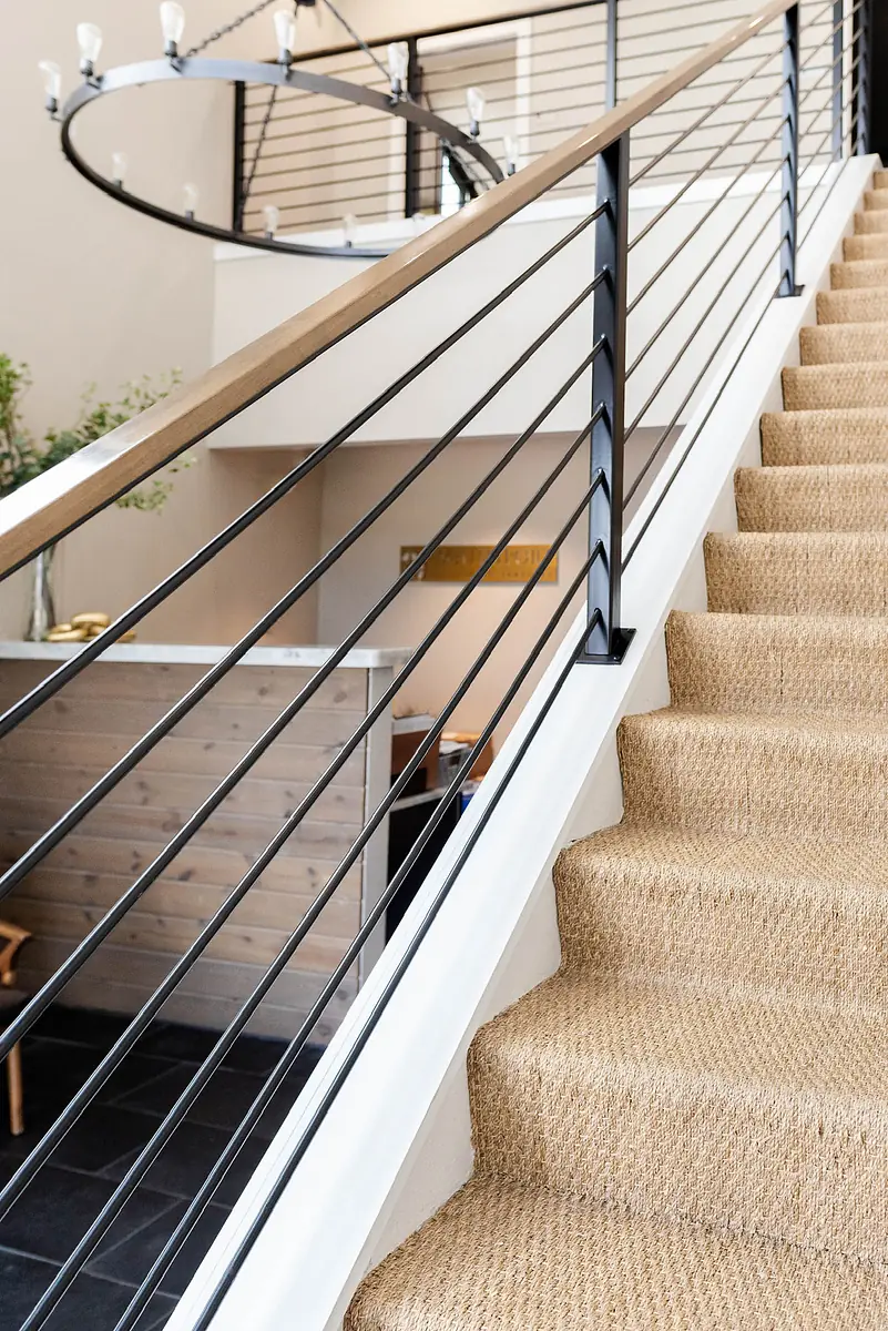 Hallway with staircase, beige carpet, black railings, and wooden handrail alongside a small seating area.