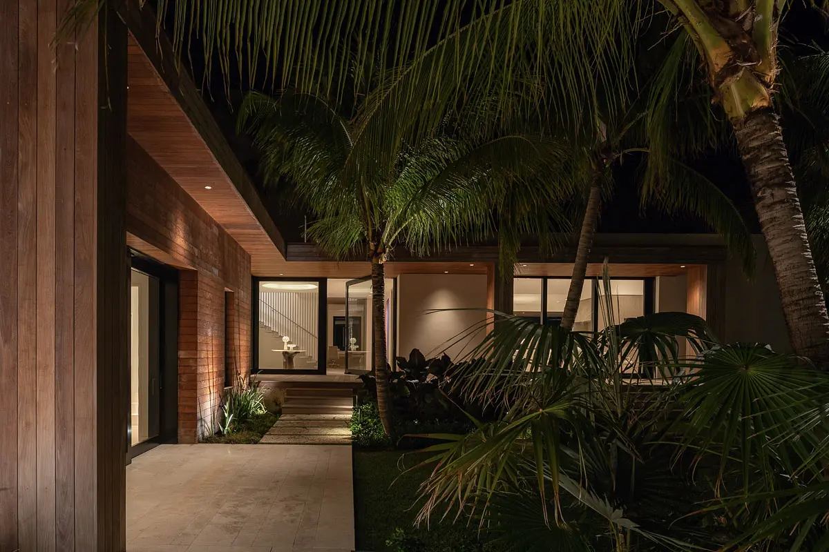 Exterior front of modern house with wood and glass panels, palm trees, stone pathway, and illuminated entrance.