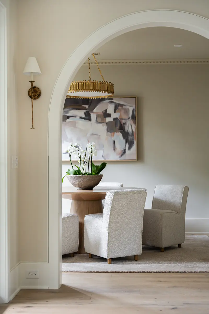 Dining room with round wooden table, two upholstered chairs, abstract painting, and greenery.