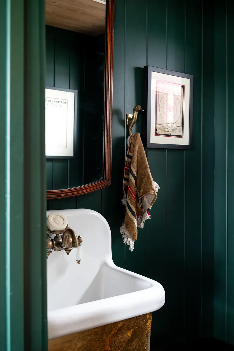 Powder room with pedestal sink, wooden framed mirror, green paneling, artwork, and gold towel holder.