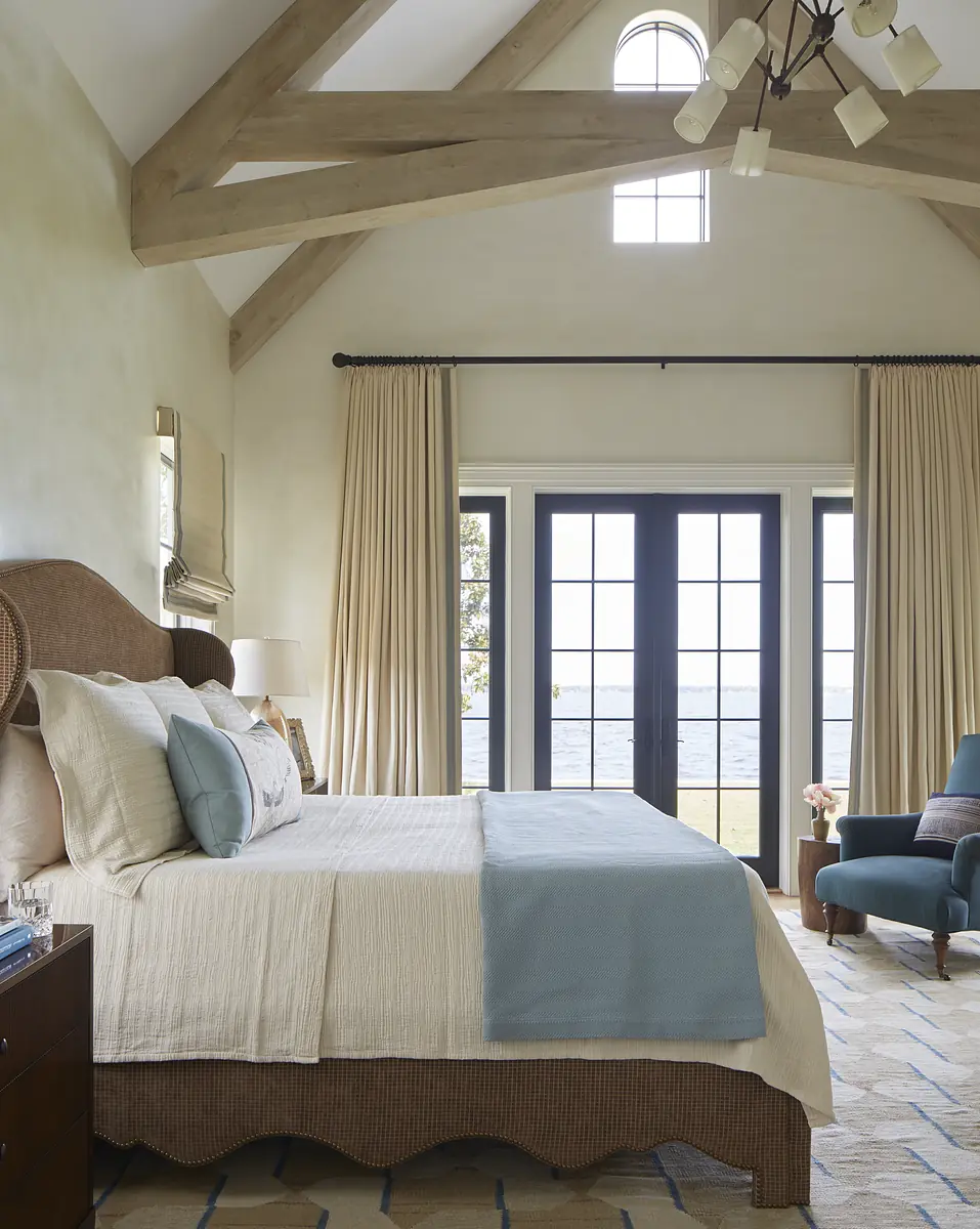 Bedroom with large bed, nightstands, blue armchair, vaulted ceilings, and double doors overlooking water