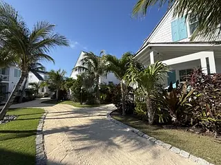 Exterior view with sandy pathway, palm trees, colorful shrubs, and houses with white siding and blue shutters.