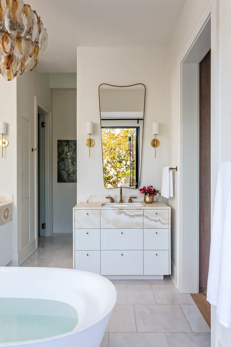 Bathroom with freestanding white bathtub, marble vanity, mirror, two sconces, and large white tiles.
