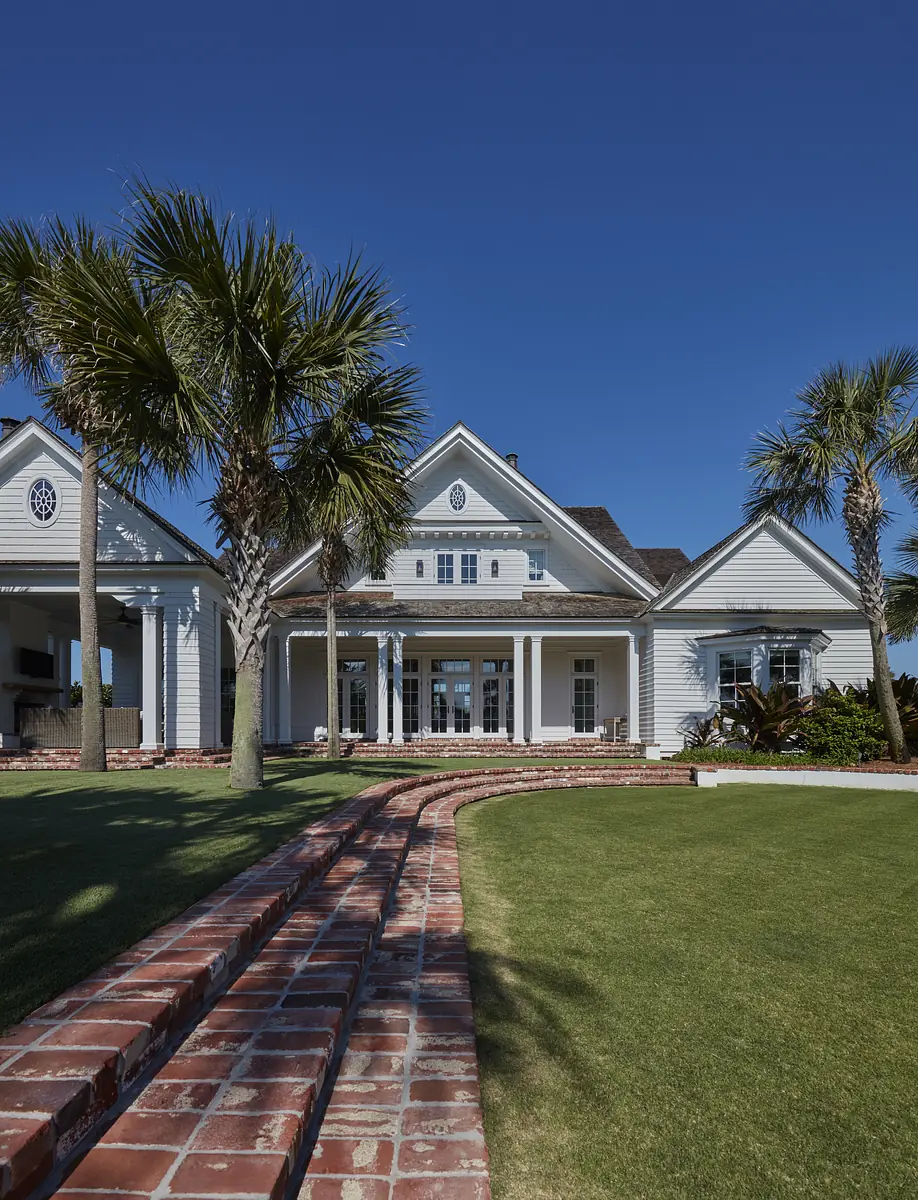 Front exterior of a house with white facade, large windows, brick stairs, and palm trees.