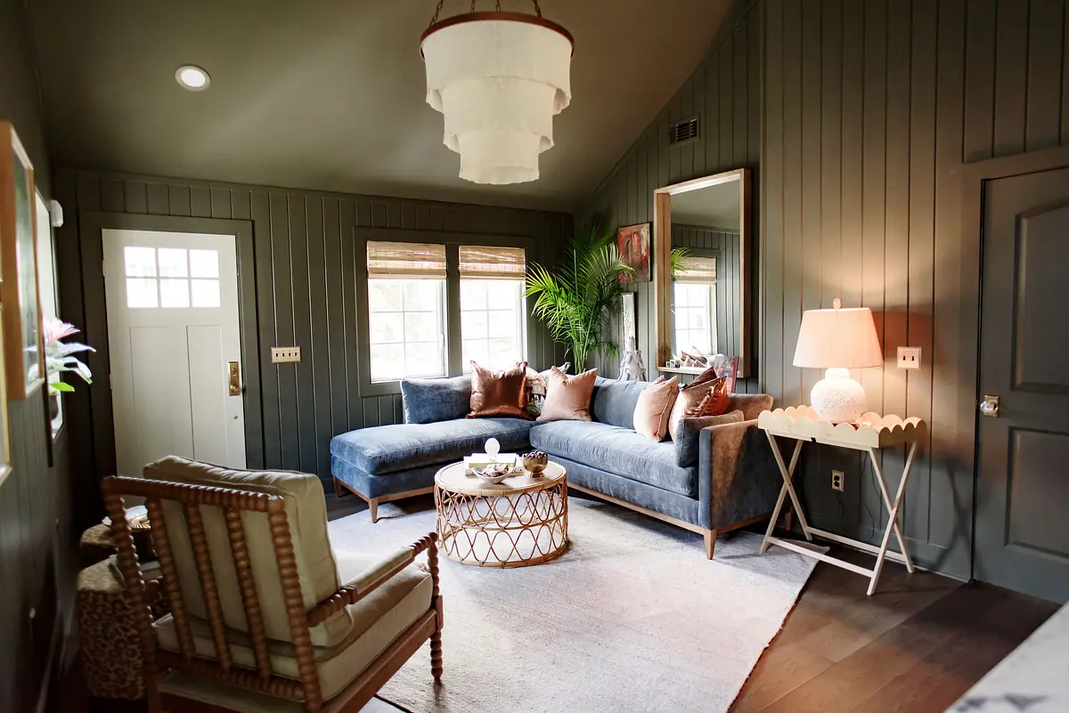 Living room with blue sectional sofa, coffee table, armchair, mirror, and large windows with blinds.