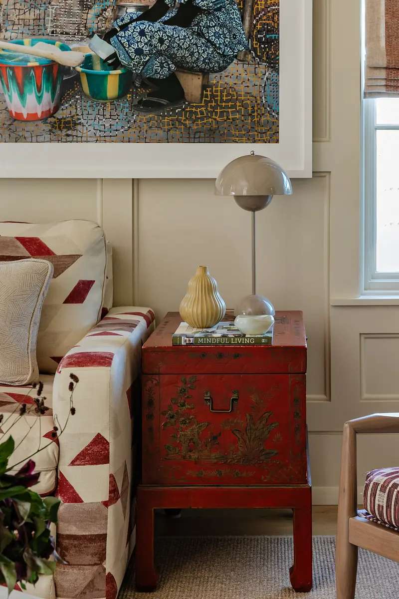 Living room with patterned sofa, red side table, table lamp, decorative vase, book, and wall art