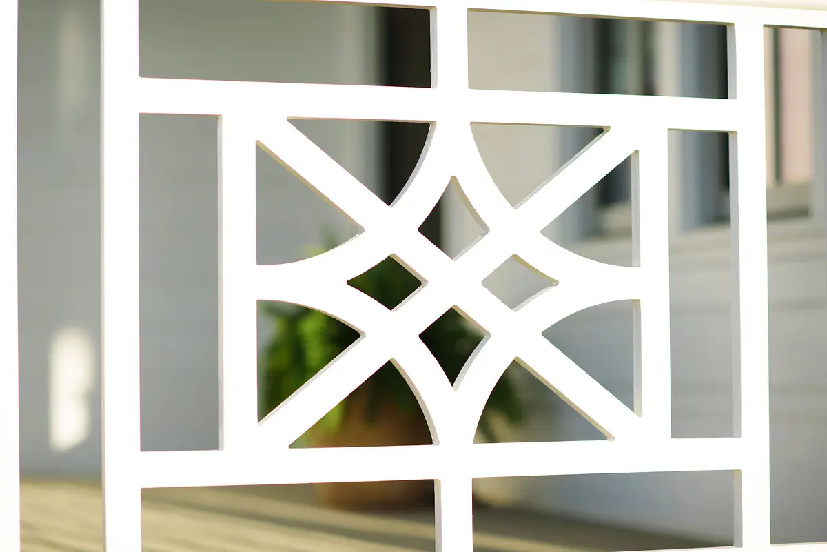 Close-up of a white decorative railing with geometric patterns and a blurred green plant in the background.
