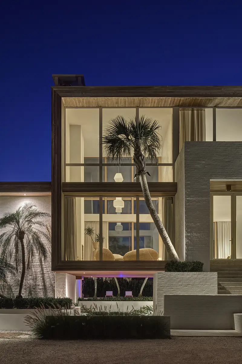 Modern house exterior with large glass windows, wooden accents, palm trees, and landscaped areas.