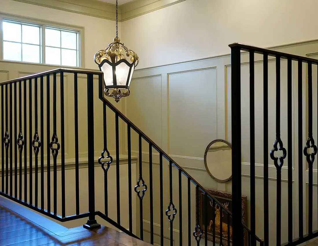 Hallway with black railing, decorative chandelier, wall mirror, and paneled walls with natural light from window