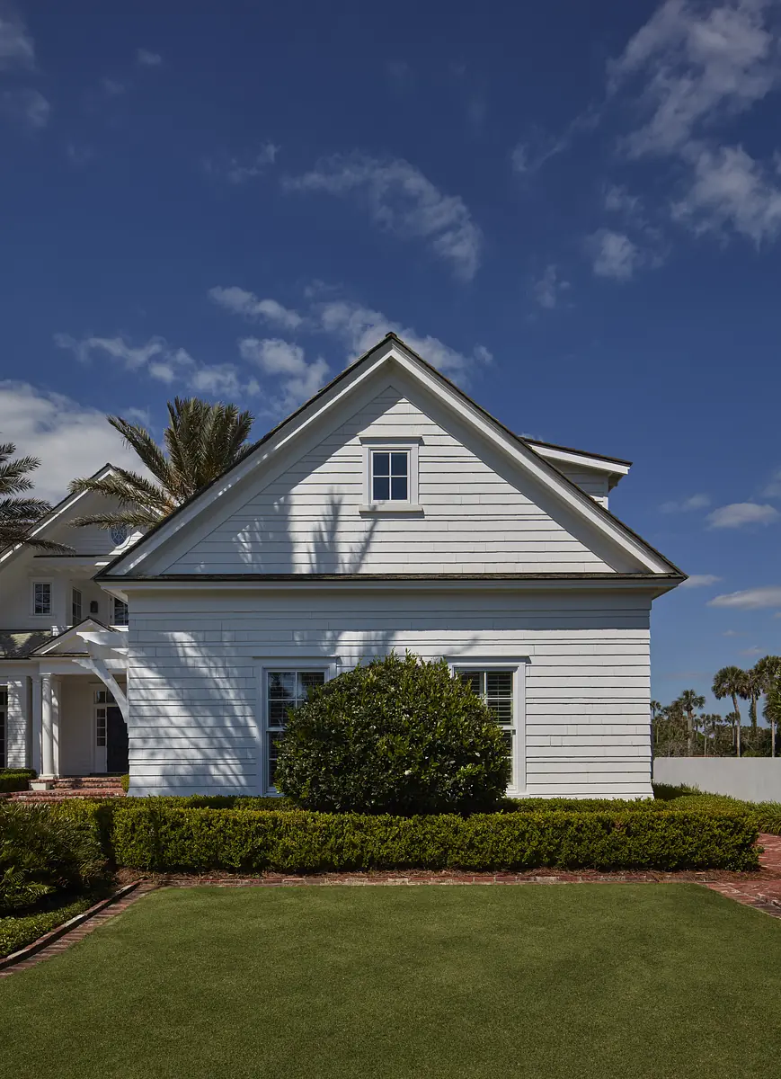 Exterior of a house with white siding, gable roof, window, bush, lawn, and palm trees against a blue sky