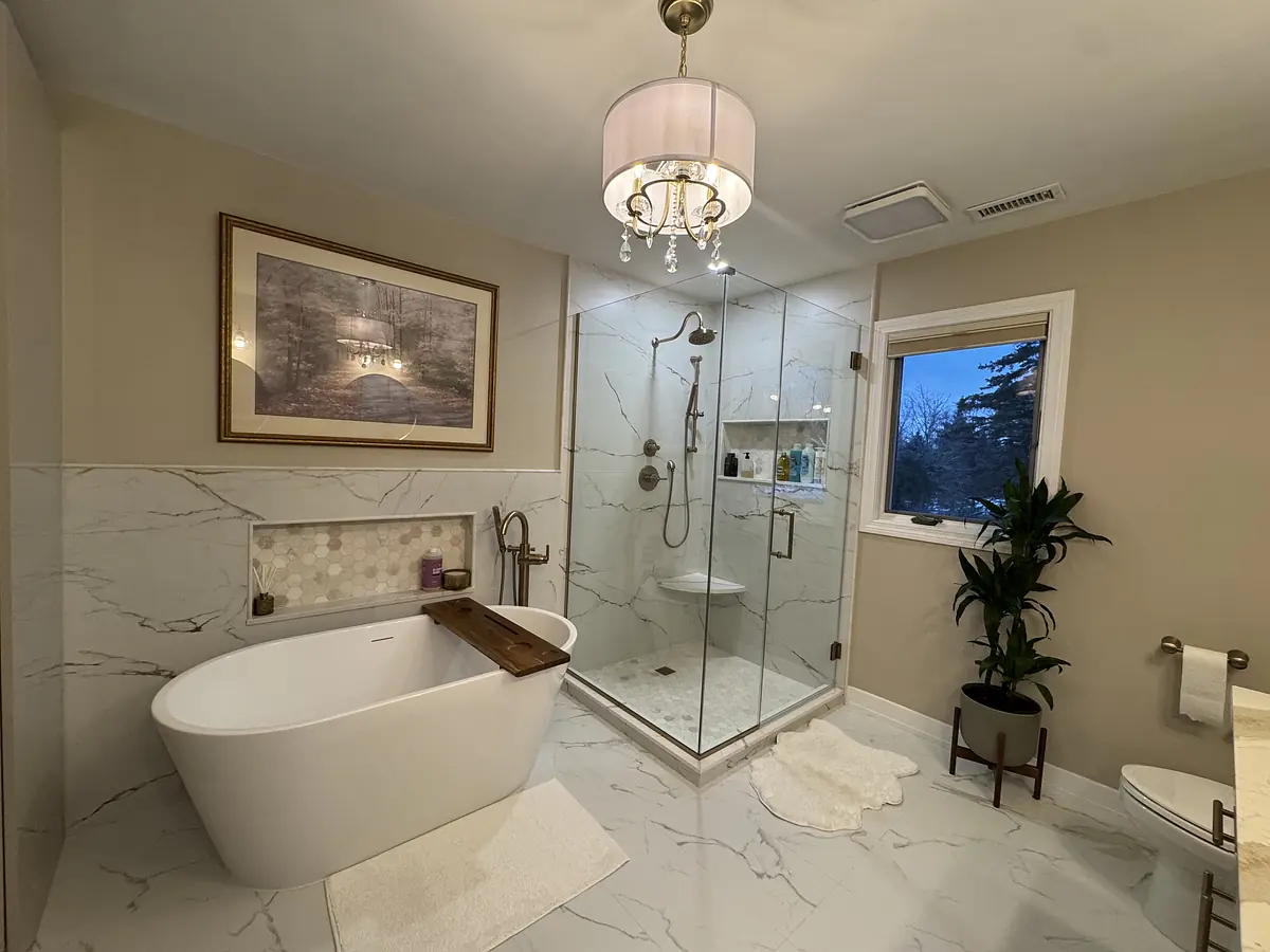 Bathroom with freestanding tub, glass shower, marble tiles, artwork, plant, and natural light from a window.