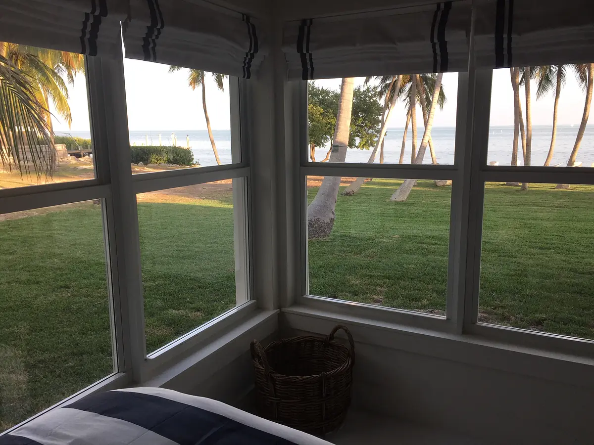 Corner view with large windows, navy-striped blinds, grassy area, and palm trees by the water.