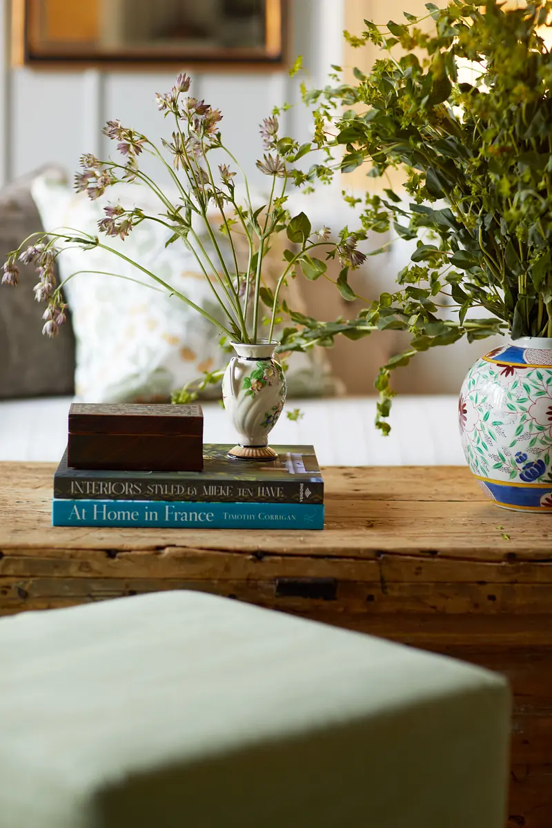 Living room area with wooden table, stacked books, vases, green stool, and decorative cushion.