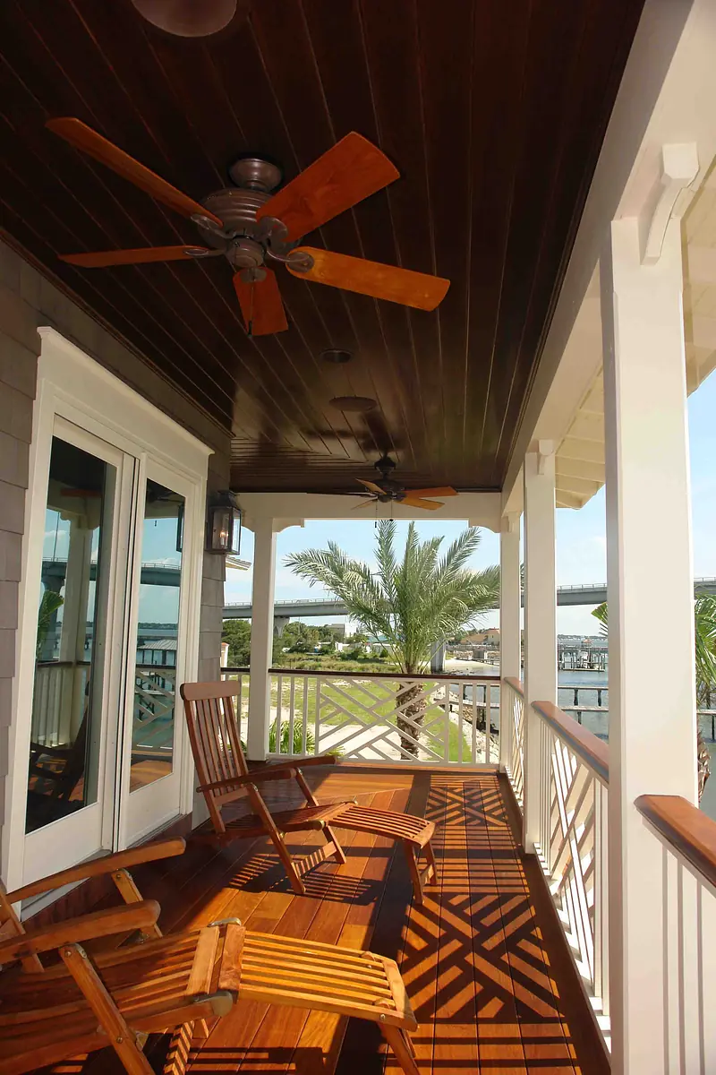 Deck with wooden flooring, rocking chairs, ceiling fan, and view of palm trees and water.
