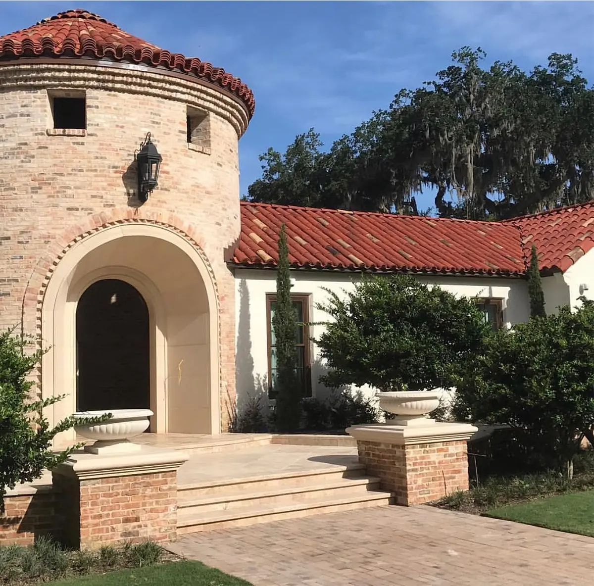 Exterior front view of house with circular tower, arched doorway, brick facade, tiled roof, and landscaping.