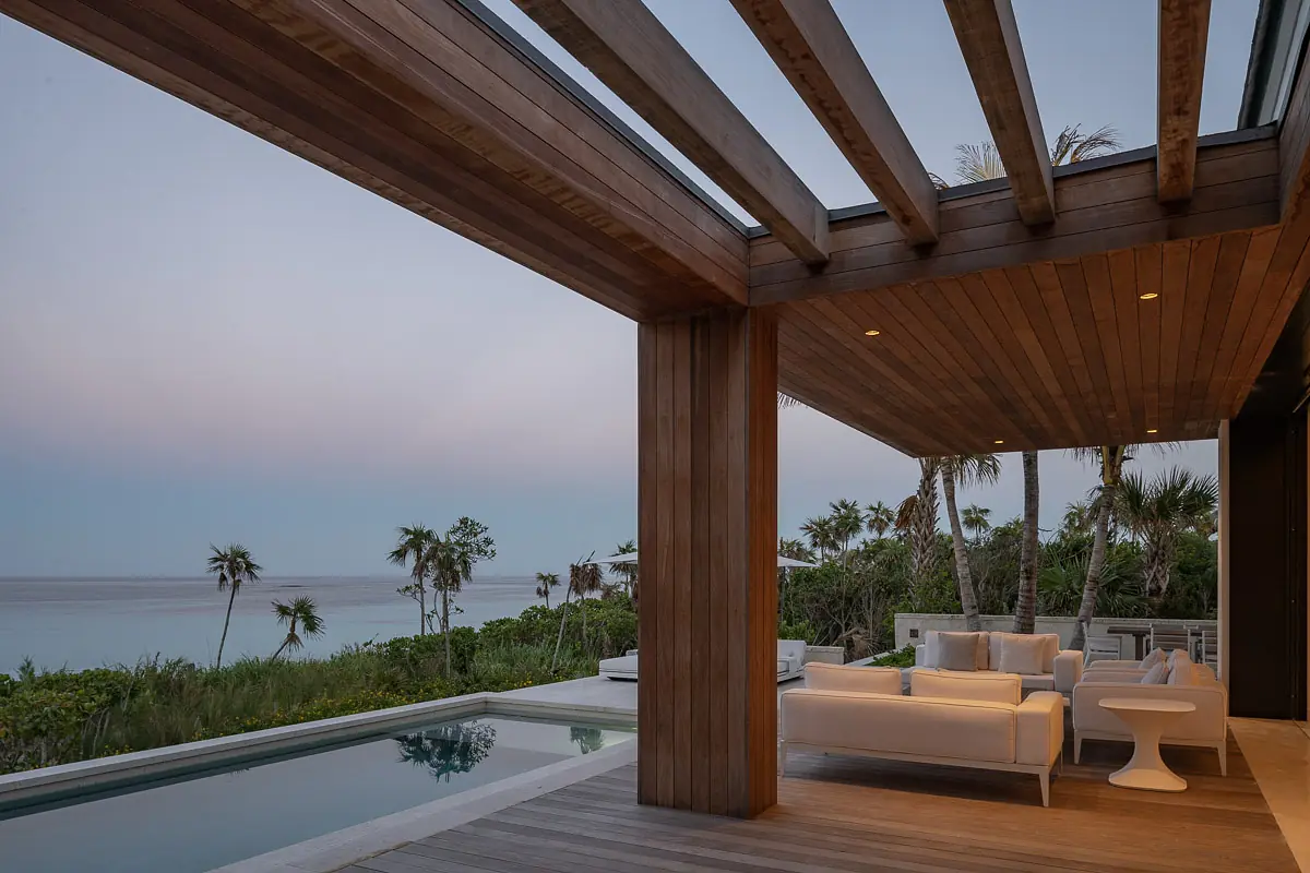 Patio with white sofa, round side table, wooden flooring, swimming pool, and greenery surrounding the area.