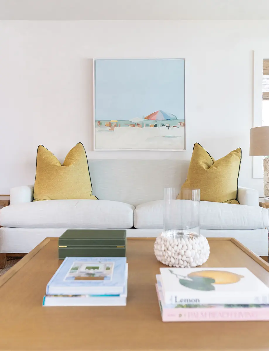 Living room with white sofa, yellow pillows, wooden coffee table, glass vase, and beach artwork.