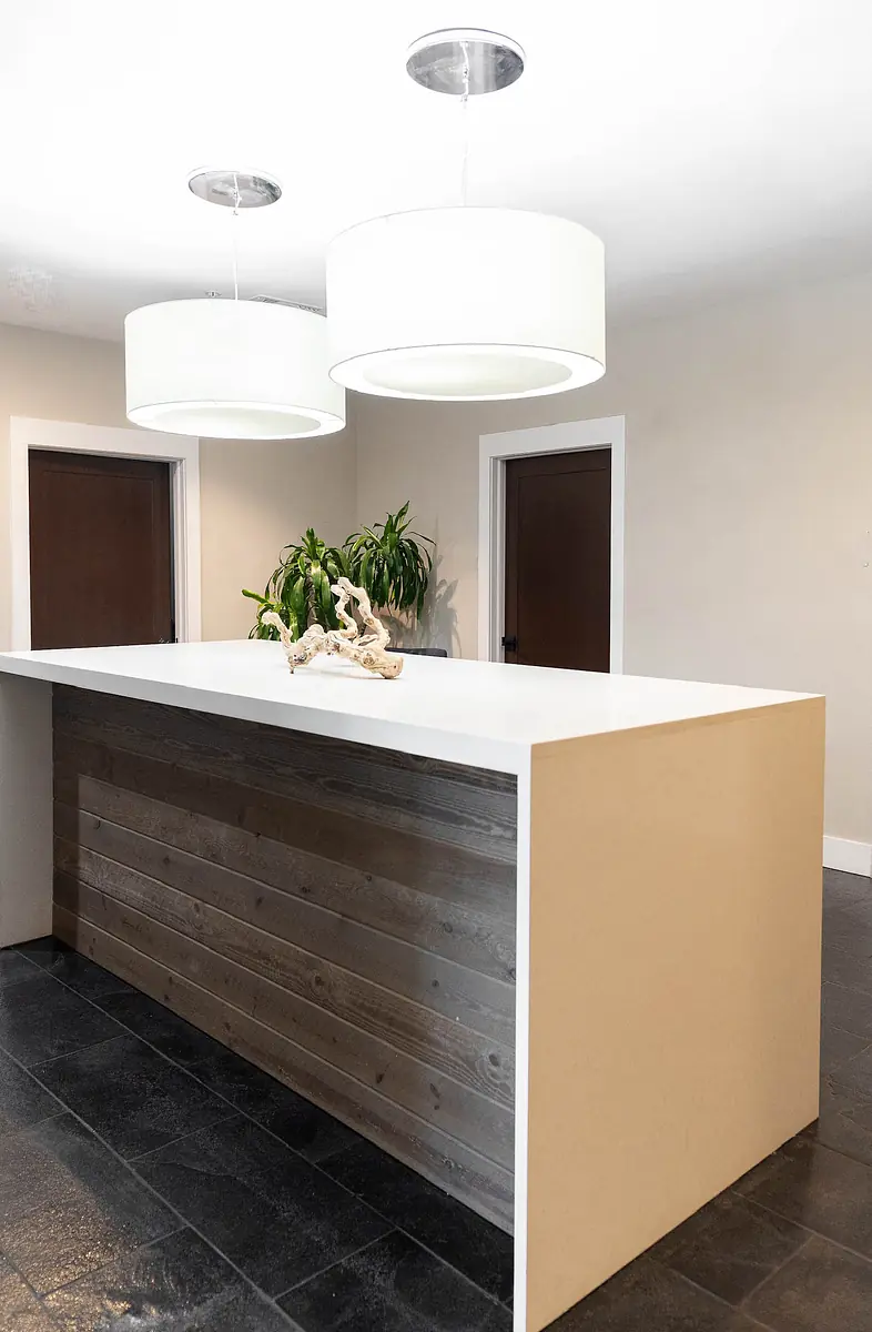 Kitchen area with large island, white countertop, wooden base, and stone floor, featuring pendant lights and plants.