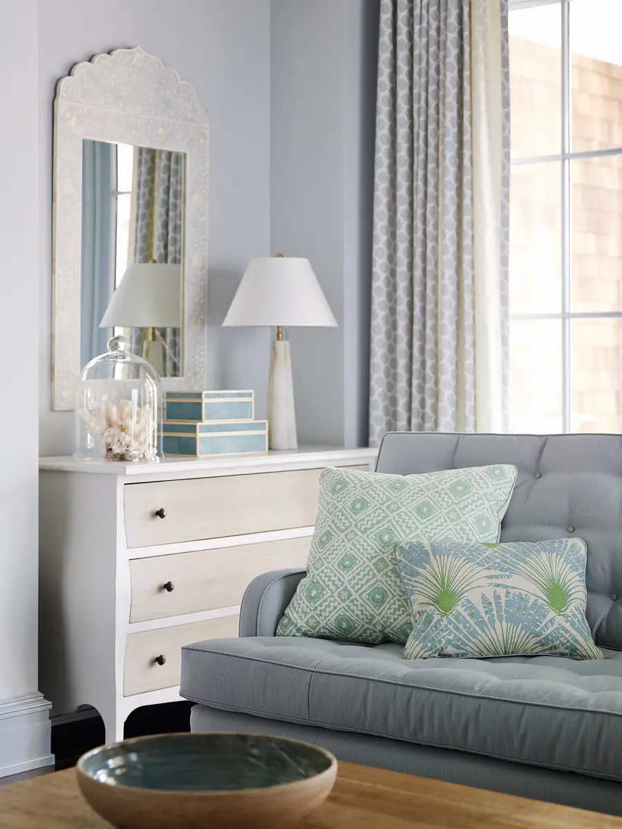 Living room with gray sofa, decorative items, dresser, and large windows with patterned curtains.