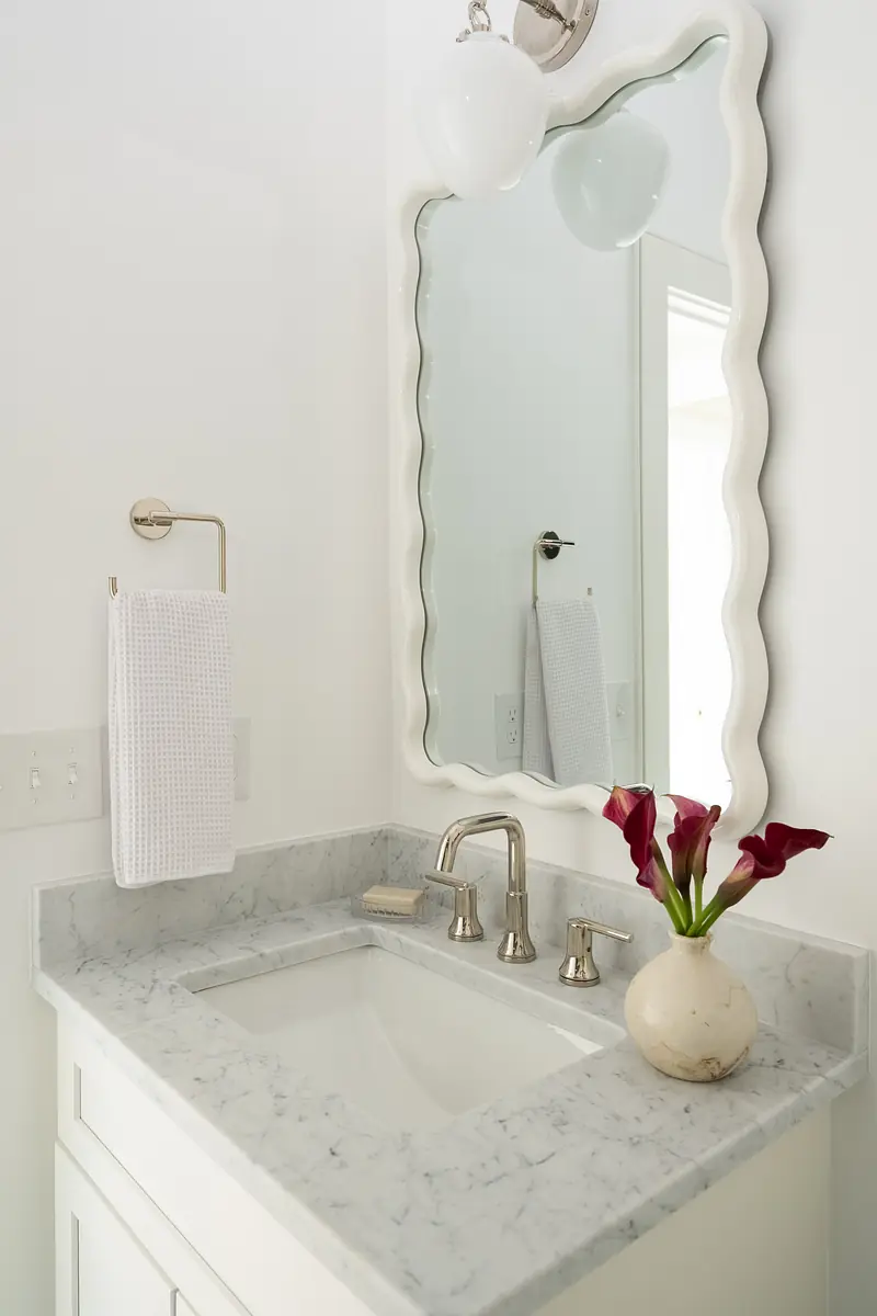 Bathroom with white vanity, marble countertop, scalloped mirror, chrome faucet, and calla lilies.