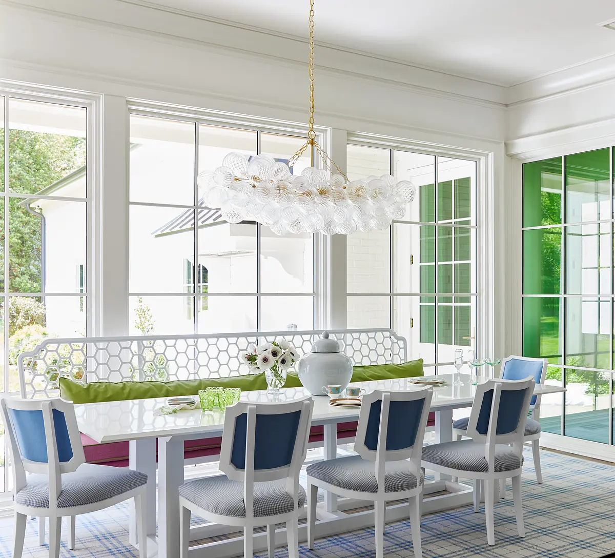 Dining room with white table, blue and white chairs, light fixture, windows, sideboard, and floral decor.