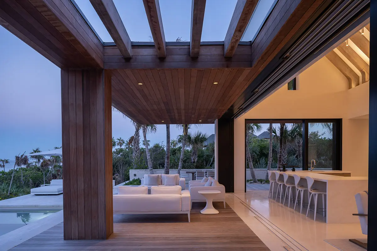 Living room with white sectional sofas, side table, bar counter, exposed beams, and large sliding glass doors.