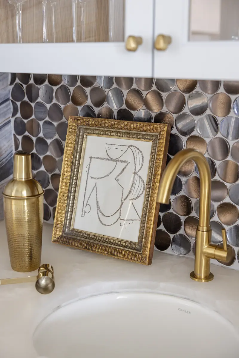 Wet bar area with gold faucet, cocktail shaker, framed artwork, and round tile backsplash.