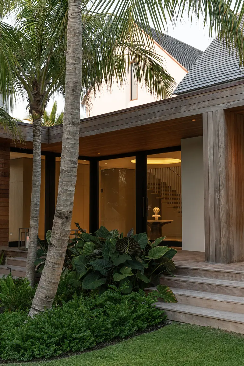 Exterior front view of a house with wooden structures, glass doors, palm trees, and green landscaping.