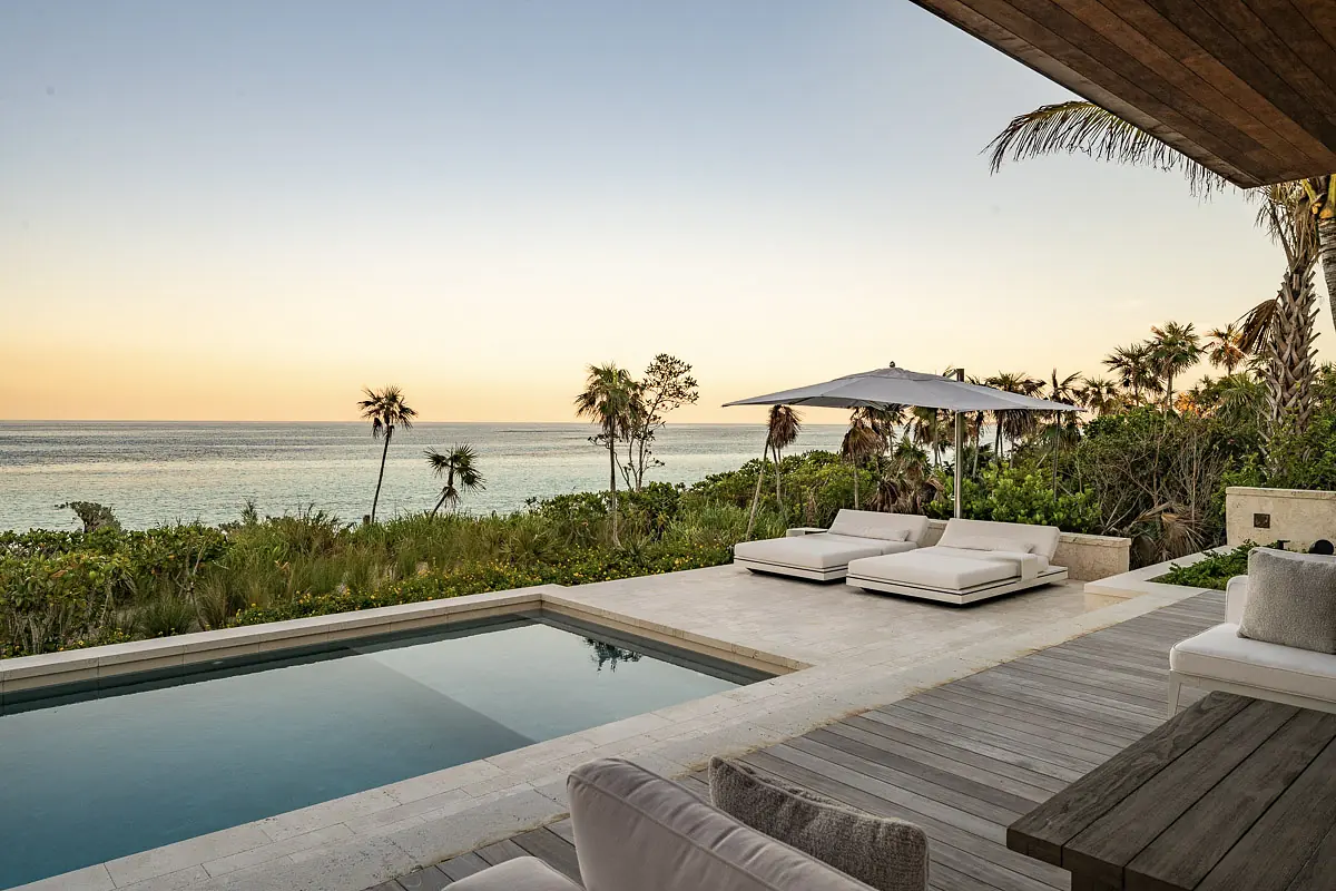 Patio with white lounge chairs, swimming pool, greenery, and a large umbrella