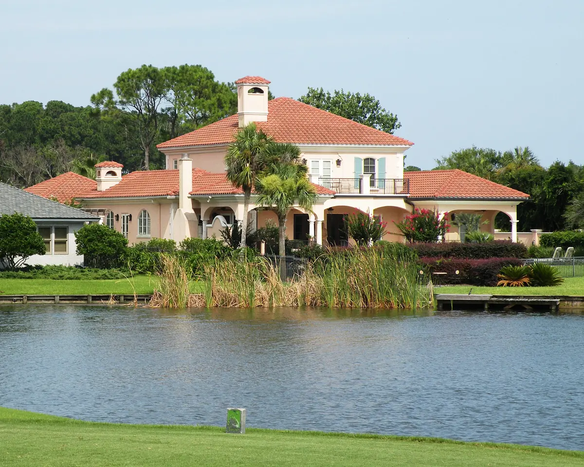 Two-story house with tile roof, balconies, windows, and covered porch, next to a pond with grass and plants.