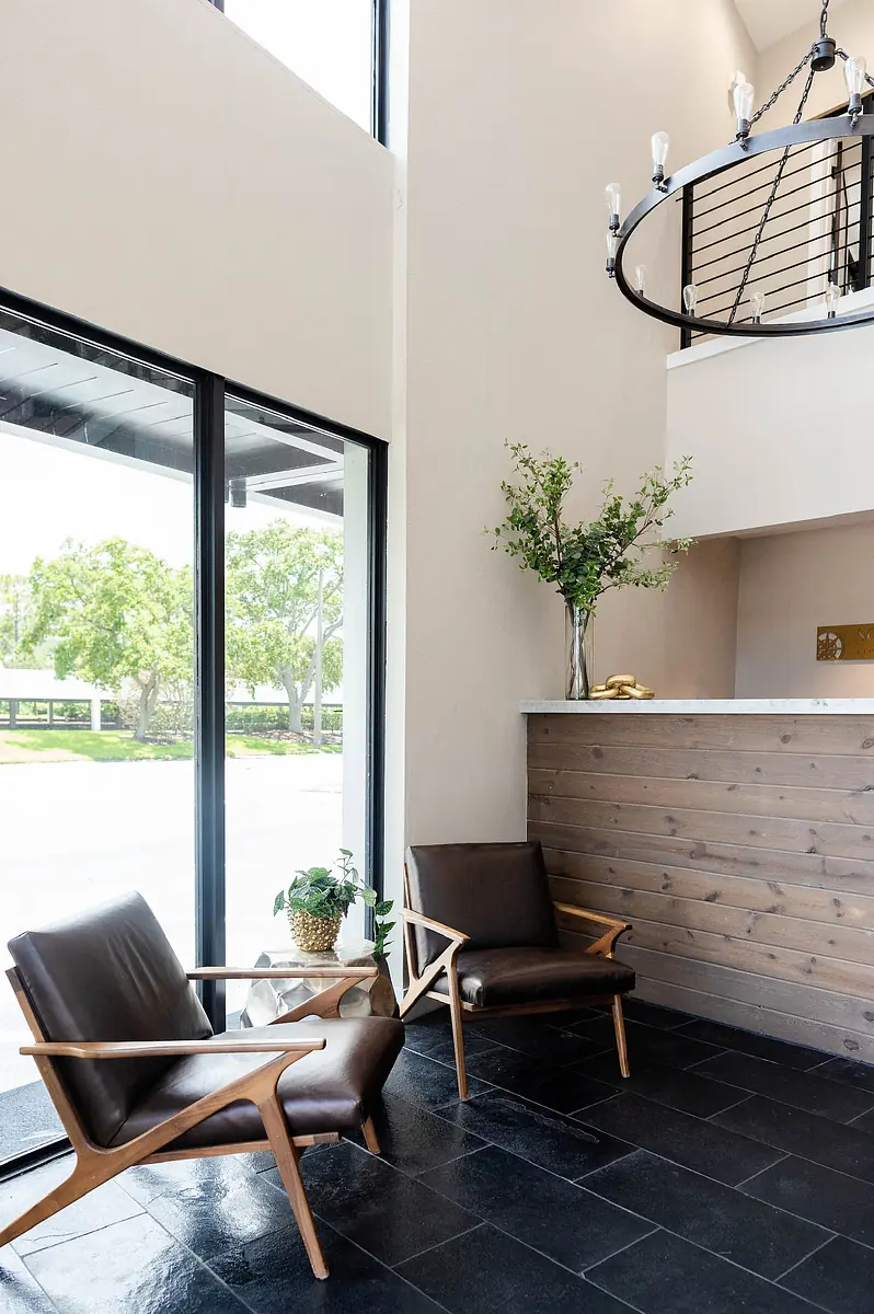 Foyer with mid-century chairs, wooden reception counter, potted plant, and large windows for natural light.