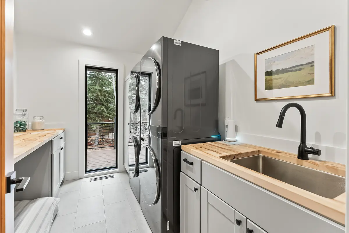 Laundry room with stacked washer and dryer, gray cabinets, wooden countertop, sink, and window.