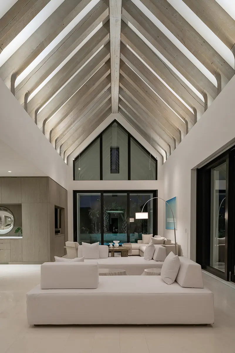Living room with white sectional sofa, light wood coffee table, large windows, and vaulted ceiling with beams.