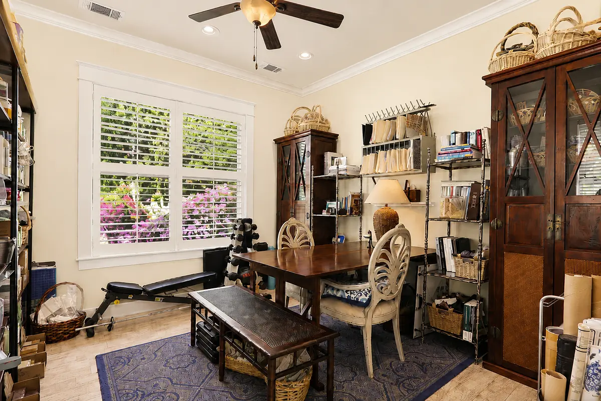 Office with wooden table, decorative chairs, shelves, rowing machine, and large window with plantation shutters.