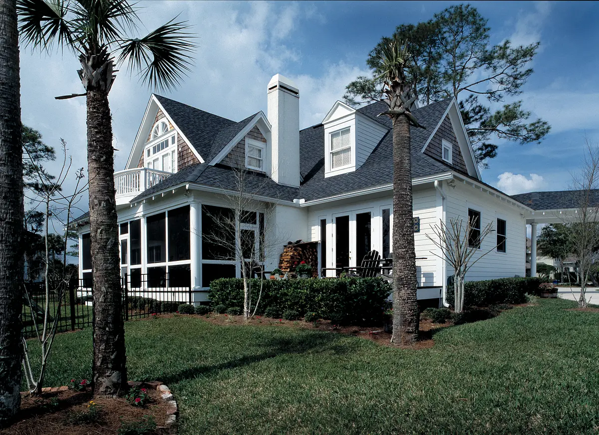 Exterior front of a house with white siding, gray roof, porch, palm trees, and trimmed hedges.