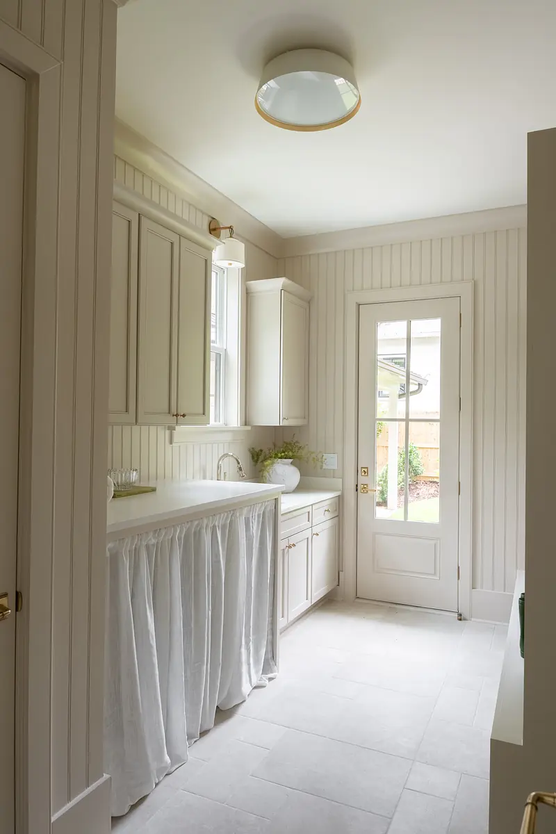 Laundry room with white cabinetry, fabric skirted countertop, beige tiled floor, and door leading outside.