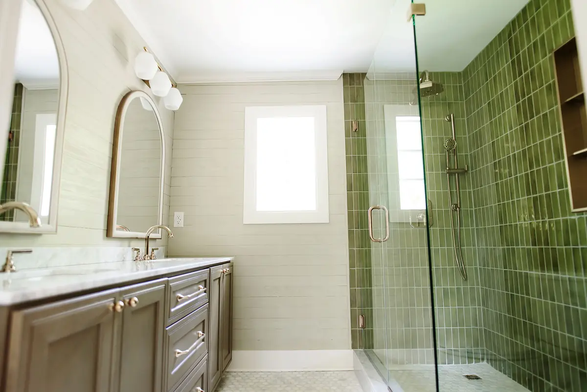 Bathroom with double vanity, green tiled shower, glass enclosure, and white window for natural light.