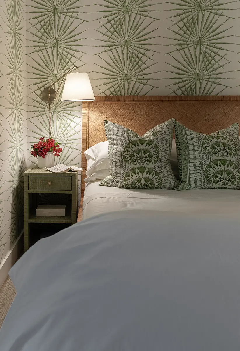 Bedroom with white bedding, patterned pillows, nightstand, lamp, and green leaf-patterned wallpaper.