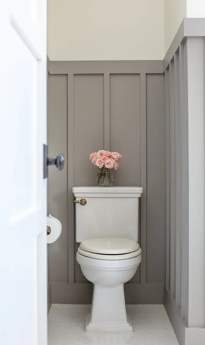 Powder room with white toilet, gray wainscoting, and vase of pink flowers.