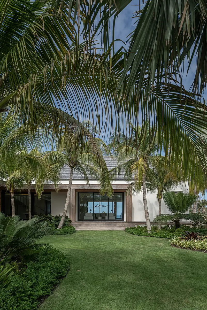Exterior front view with house, palm trees, shrubs, large windows, and a grassy area leading to a wooden deck.