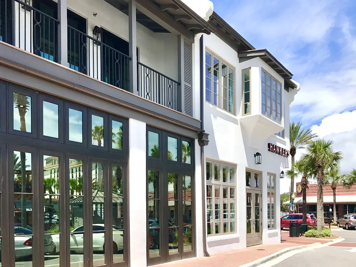 Modern commercial exterior with glass windows, balcony, potted palms, and brick pavement.