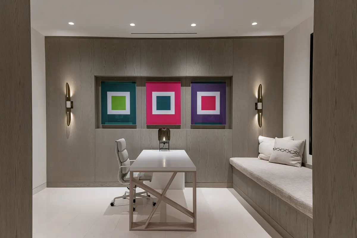 Office with light wood desk, white chair, built-in bench, and colorful framed wall art