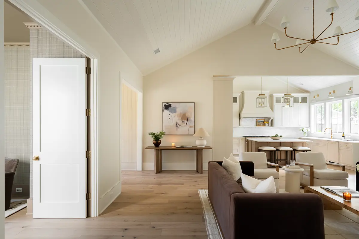 Living room with brown sofa, light wood coffee table, art piece, and sloped ceilings connecting to a kitchen.