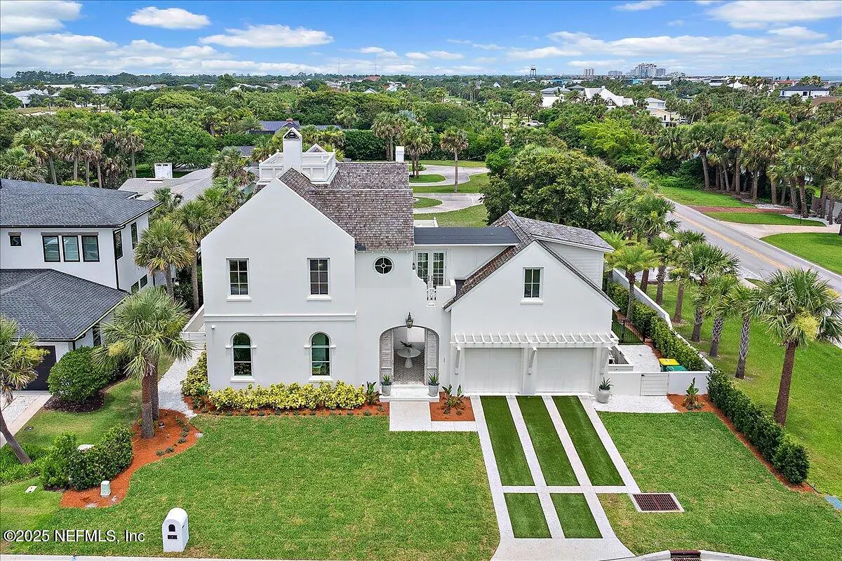 Exterior front view of a house with white stucco, dark shingles, manicured lawn, and palm trees.