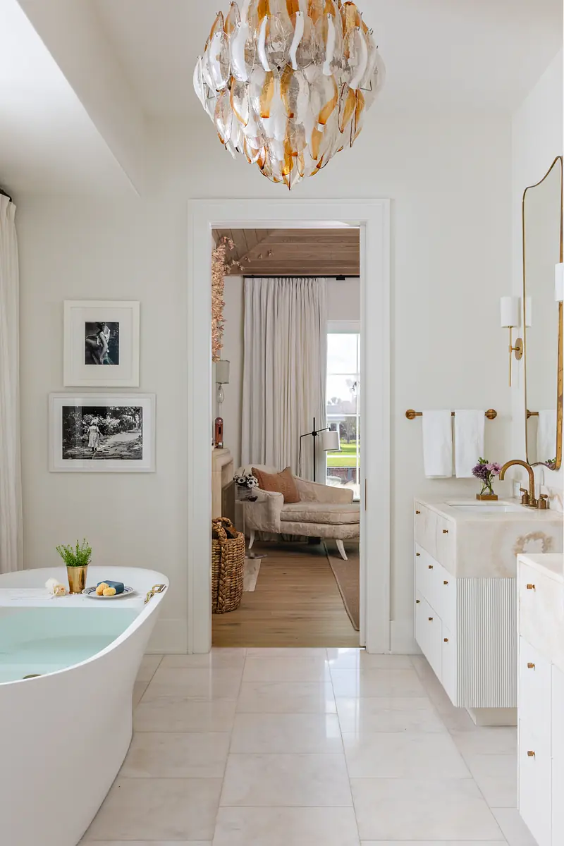 Bathroom with freestanding bathtub, white cabinetry, marble countertops, artwork, and a window with sheer curtains.