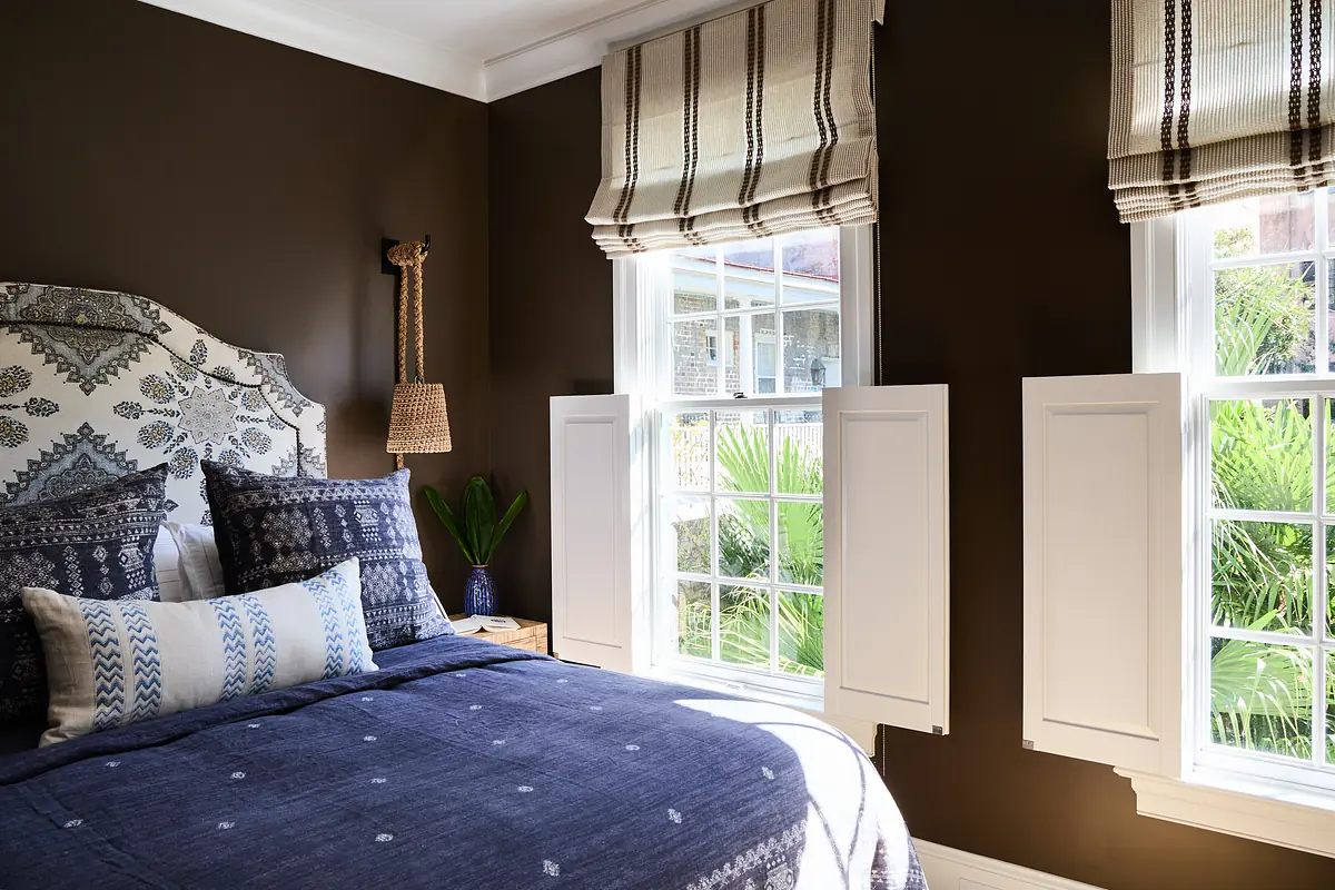 Bedroom with patterned headboard, blue bed linens, windows with shutters, and nightstand with plant.