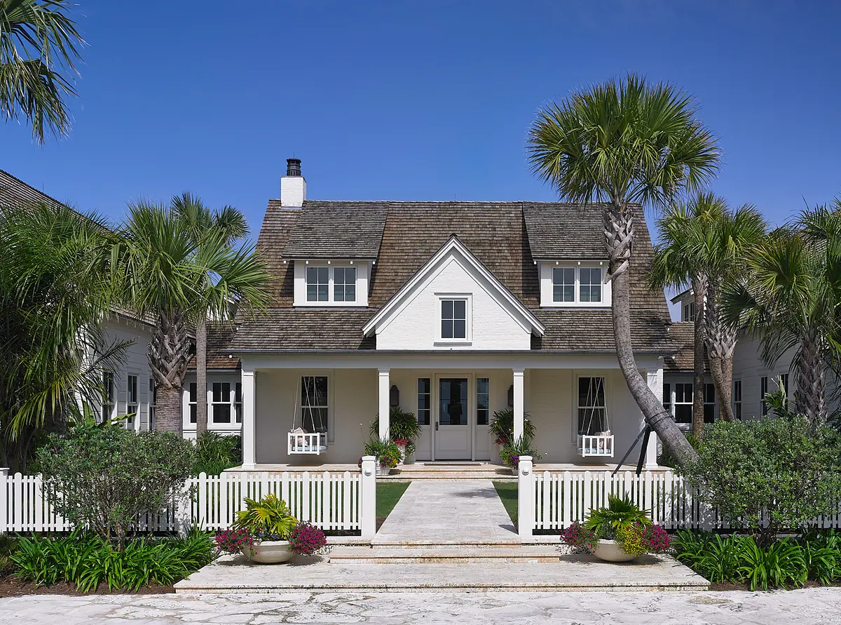 Front exterior of a house with white facade, double doors, porch, palm trees, and picket fence